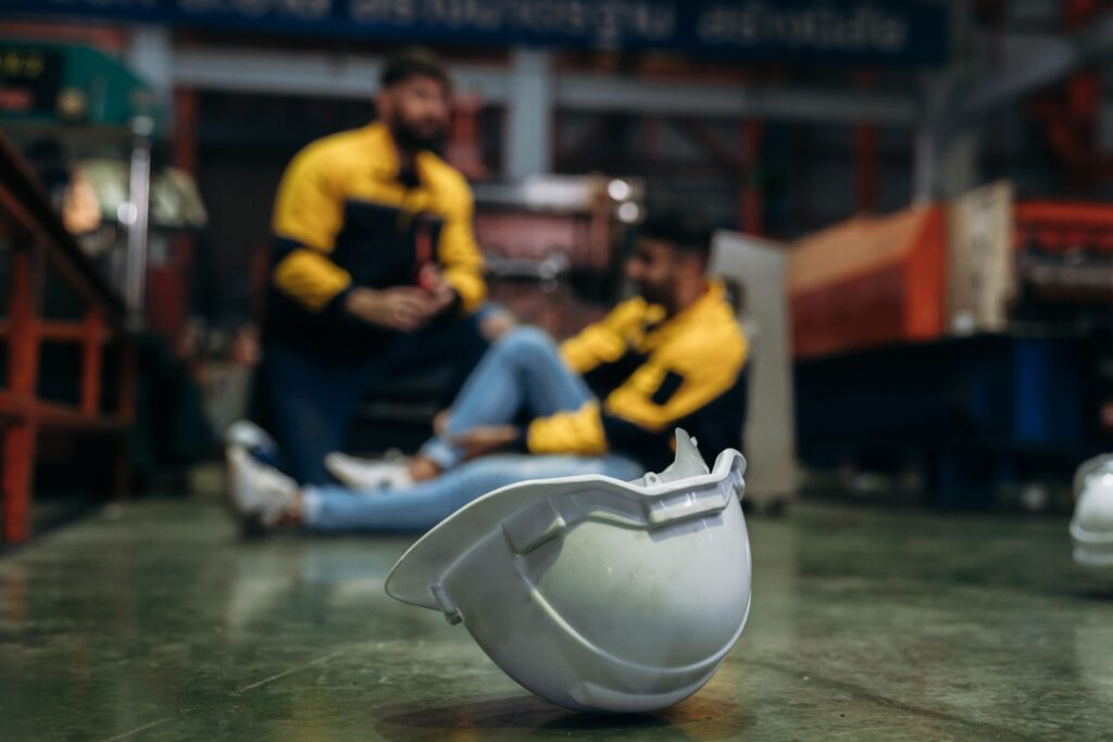 Capacete de segurança no chão de ambiente industrial, com trabalhador sendo atendido ao fundo após um possível acidente de trabalho.