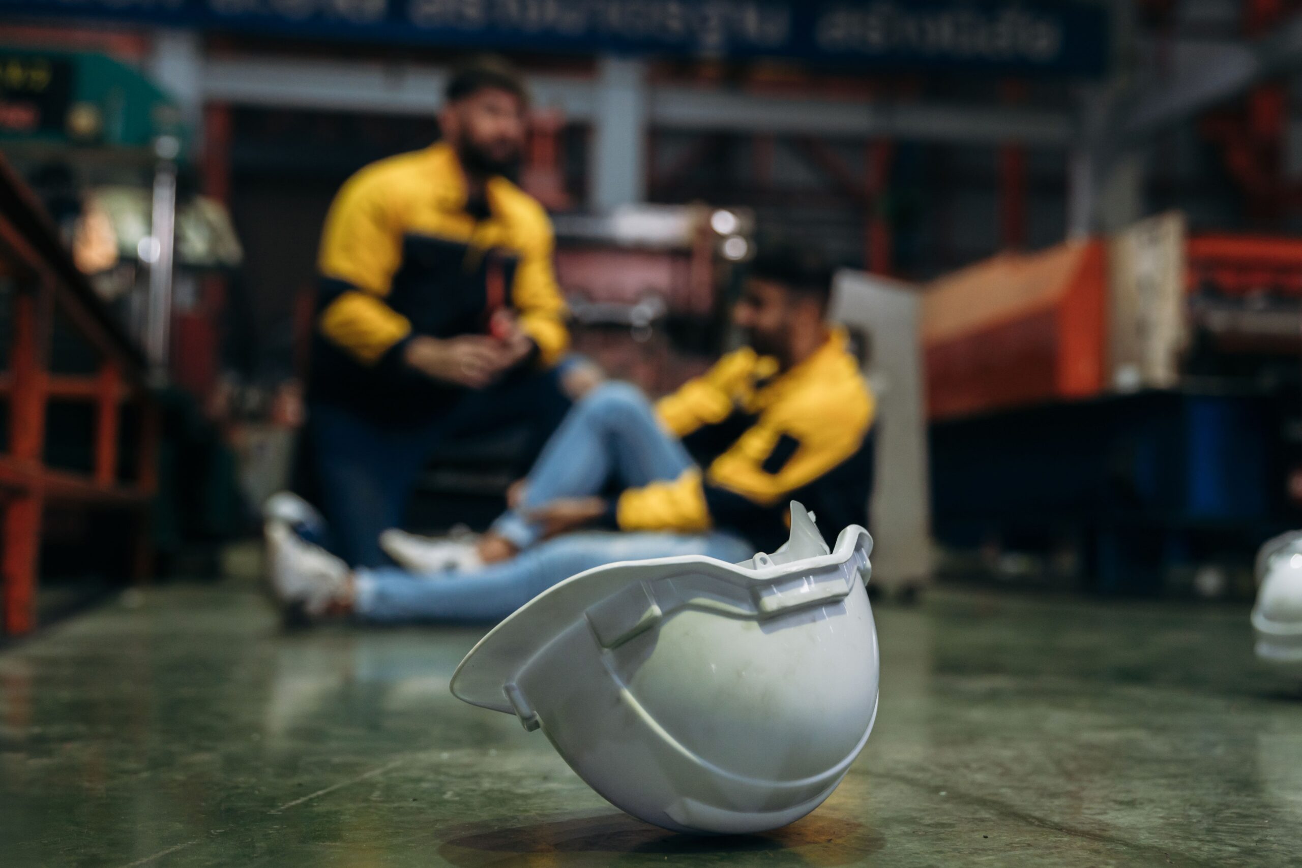 Capacete de segurança no chão de ambiente industrial, com trabalhador sendo atendido ao fundo após um possível acidente de trabalho.
